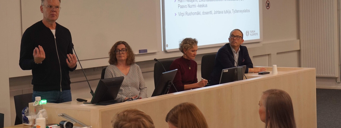 Neljä ihmistä istumassa luentosalin edessä. Yksi panelisteista puhuu.