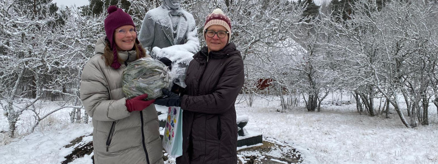 Lumisessa maisemassa kaksi naista onnitteluseppeleen kanssa. Taustalla Elias Lönnrotin patsas.