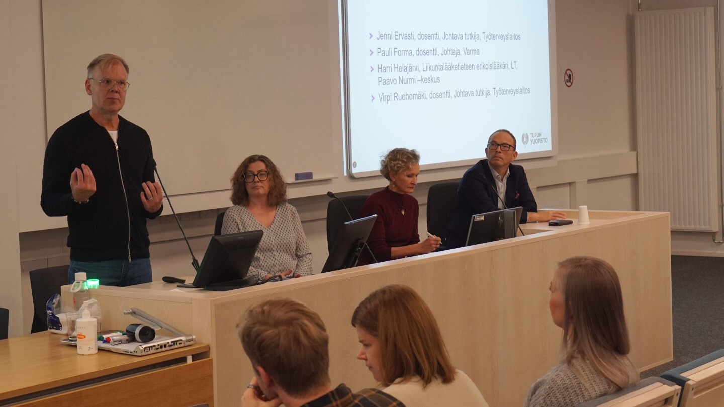 Neljä ihmistä istumassa luentosalin edessä. Yksi panelisteista puhuu.