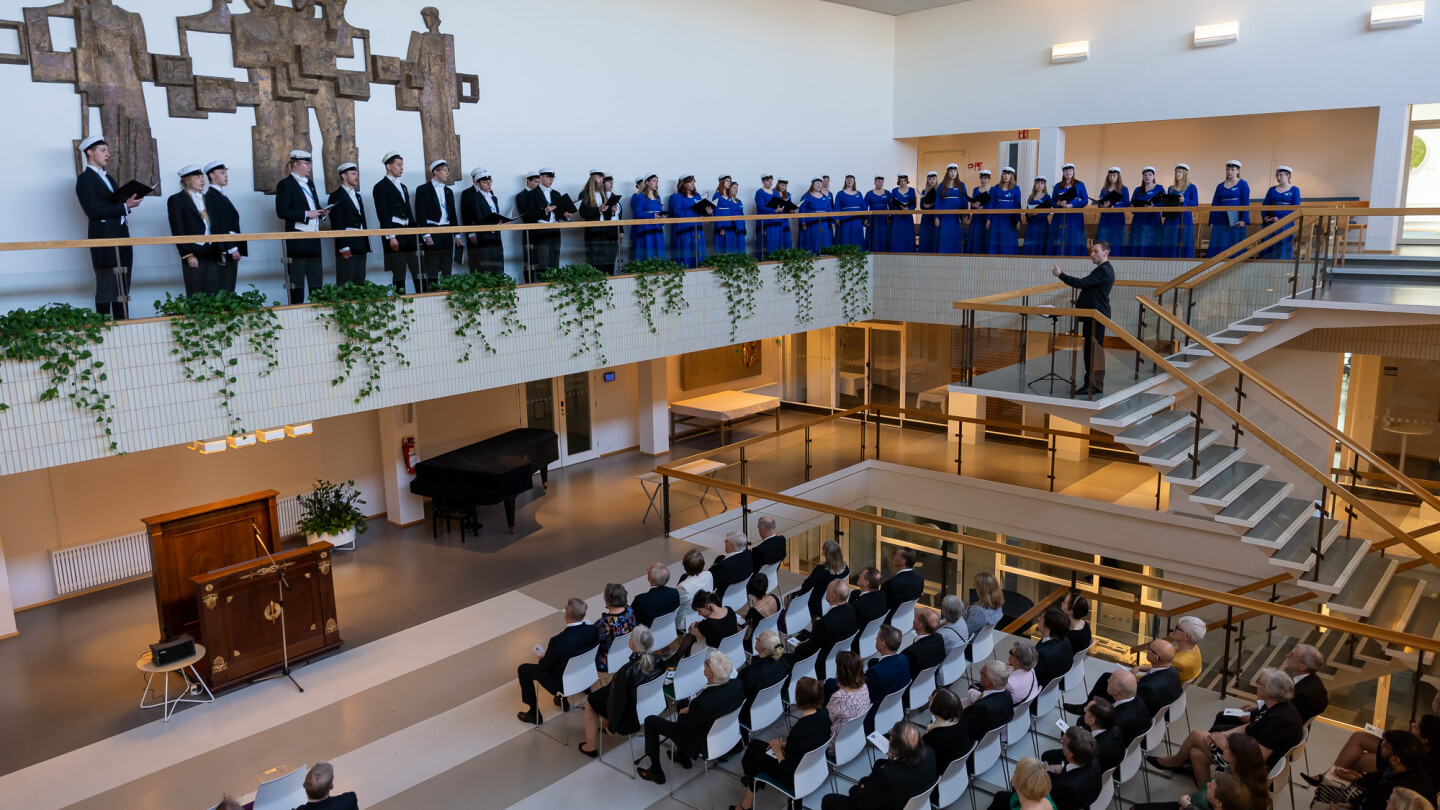 Ihmisiä istumassa Turun yliopiston päärakennuksen aulassa, portaissa kapellimestari, joka ohjaa parvella esiintyvää kuoroa.