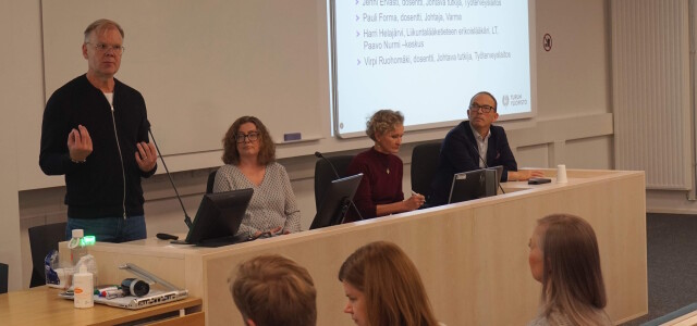 Neljä ihmistä istumassa luentosalin edessä. Yksi panelisteista puhuu.