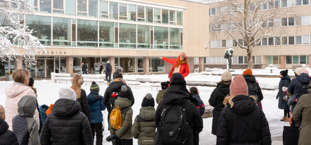 A woman in a red coat is guiding a group of children on a snowy courtyard.