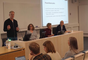 Neljä ihmistä istumassa luentosalin edessä. Yksi panelisteista puhuu.