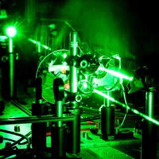 Pimeässä laboratoriossa kuvattu lähikuva vihreästä lasersäteestä läpäisemässä optiikka-laitteistossa näkyviä komponentteja